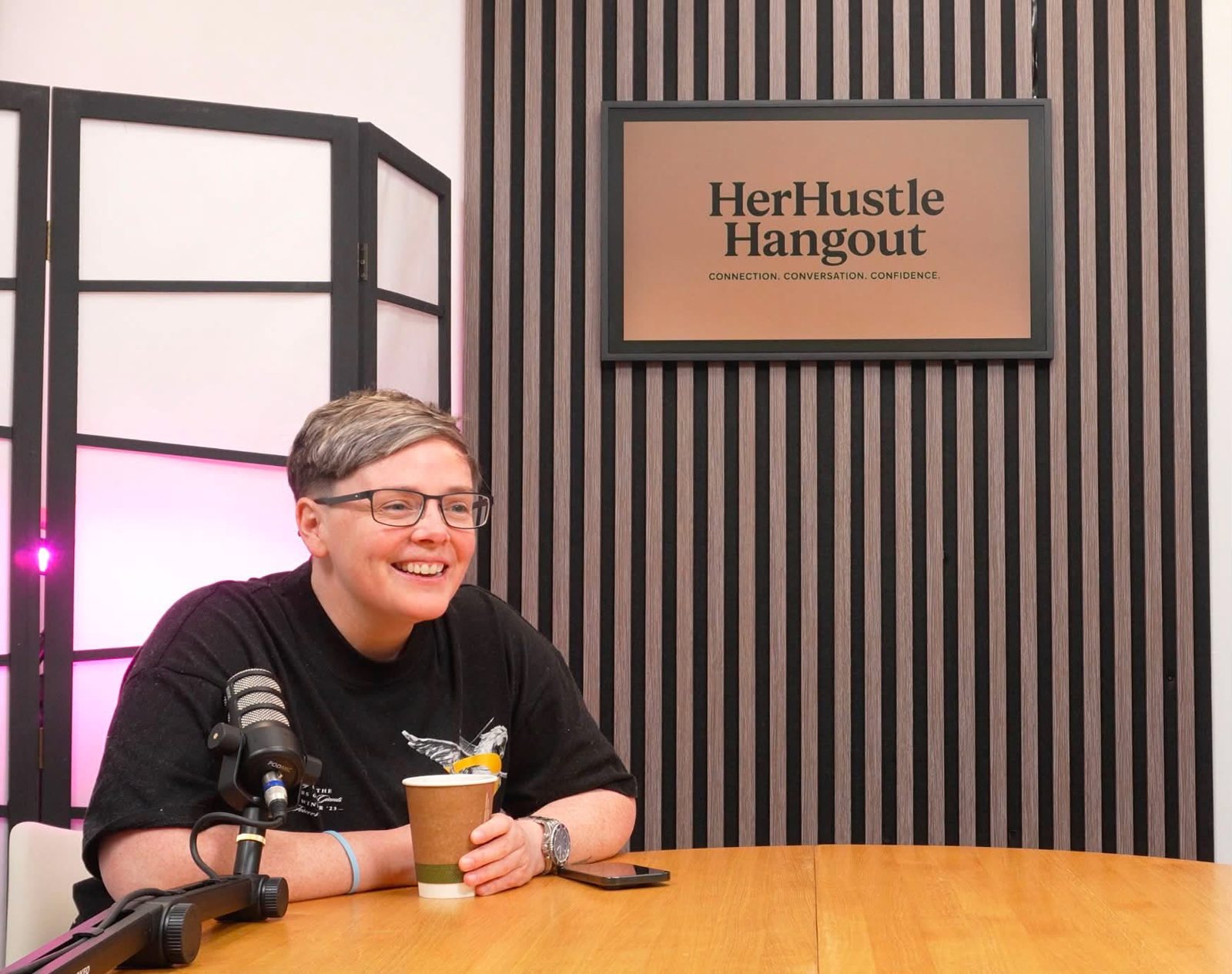 Alex Brown laughing at a table with a microphone and a coffee, in front of a HerHustle Hangout sign.