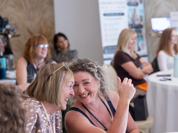 Two women laughing together, heads close, at a HerHustle event.